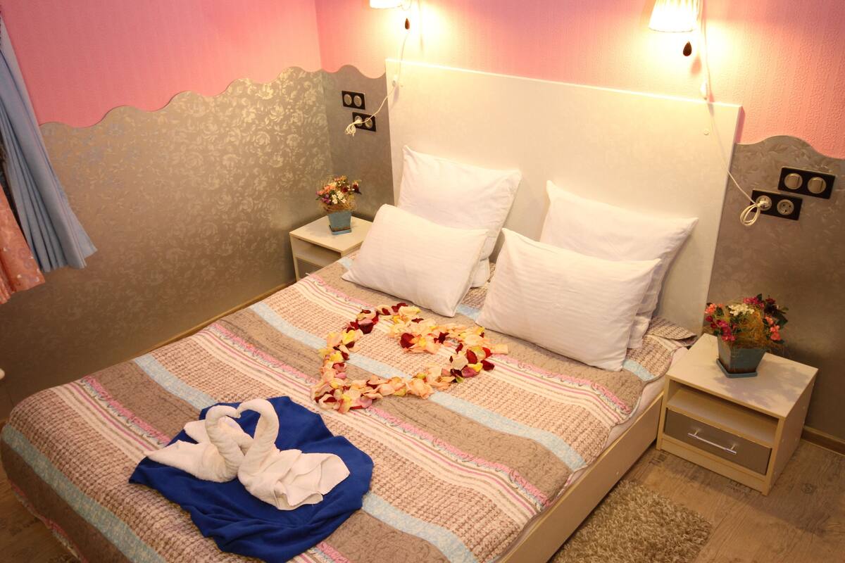 Siististi pedattu sänky, jonka päällä on sydämenmuotoinen kukka-asetelma, sininen pyyhe ja valkoinen kylpytakki. Sängyn vieressä on kaksi yöpöytää, joissa on lamput, ja toisessa niistä on pieni maljakko kukkia.