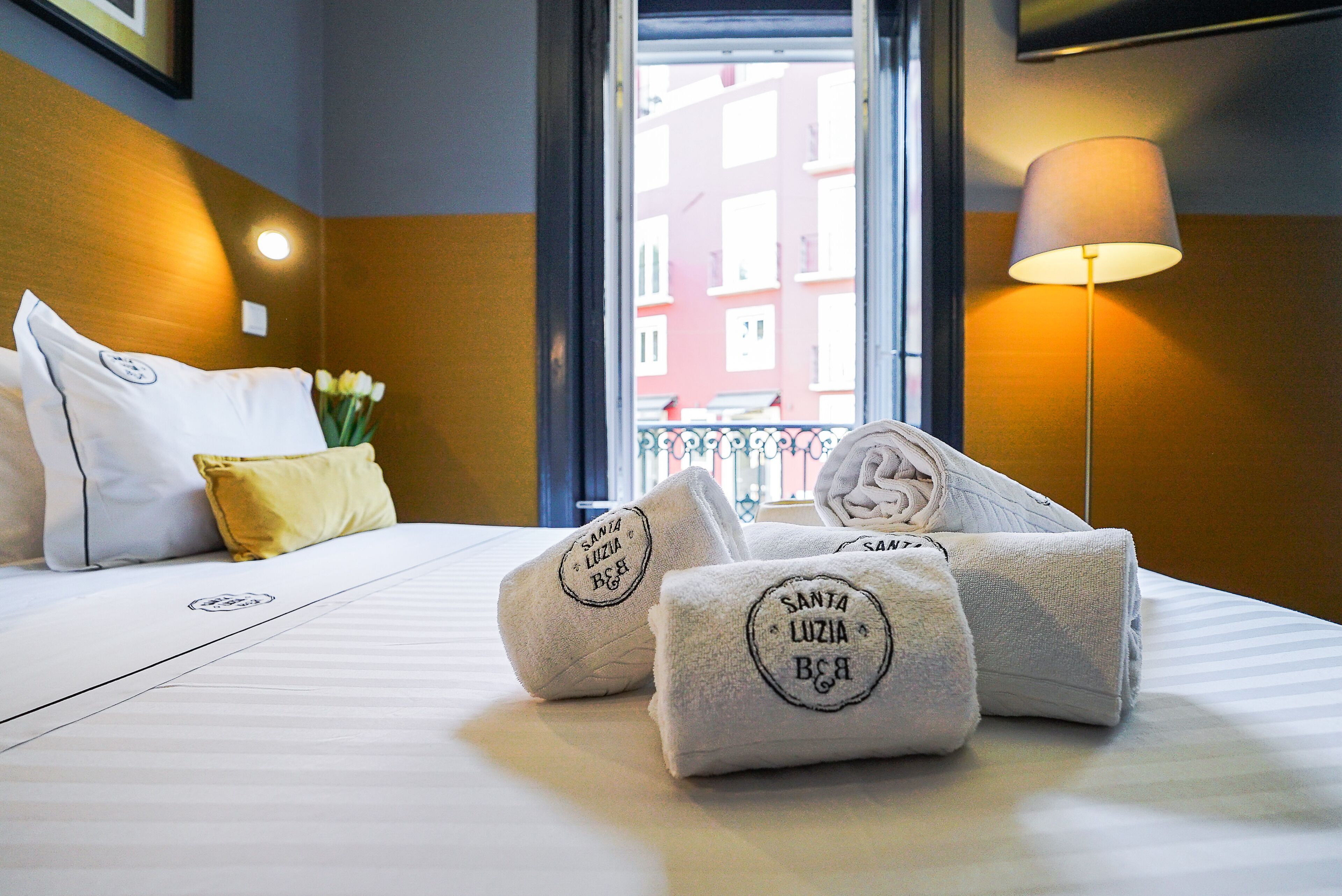 A hotel room with a neatly made bed, a window with a view of buildings, a lamp, and towels with a logo.