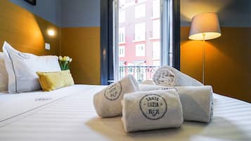 A hotel room with a neatly made bed, a window with a view of buildings, a lamp, and towels with a logo.