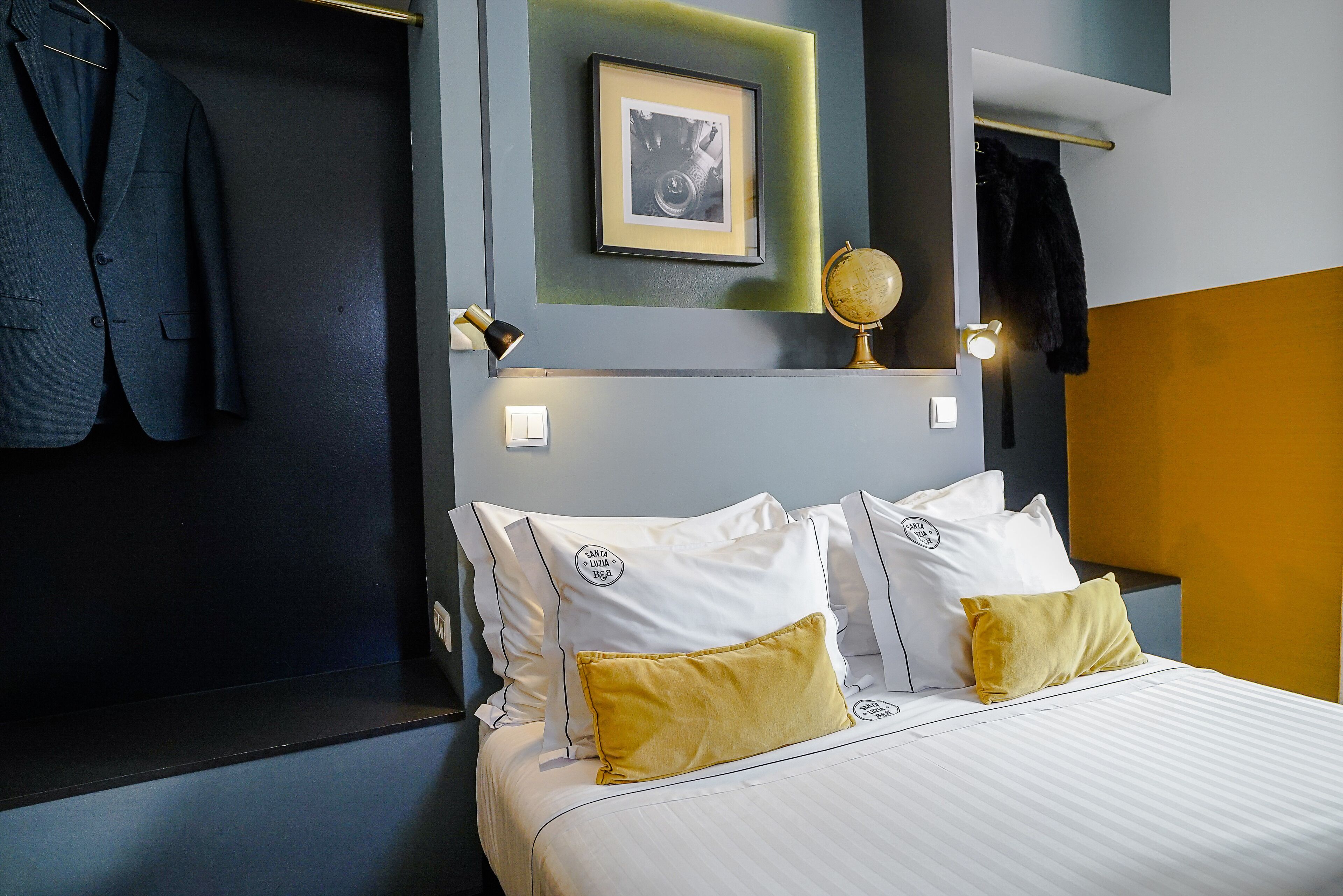 A neatly made bed with white linens and two yellow pillows, a framed picture on the wall, and a suit hanging on the wall.