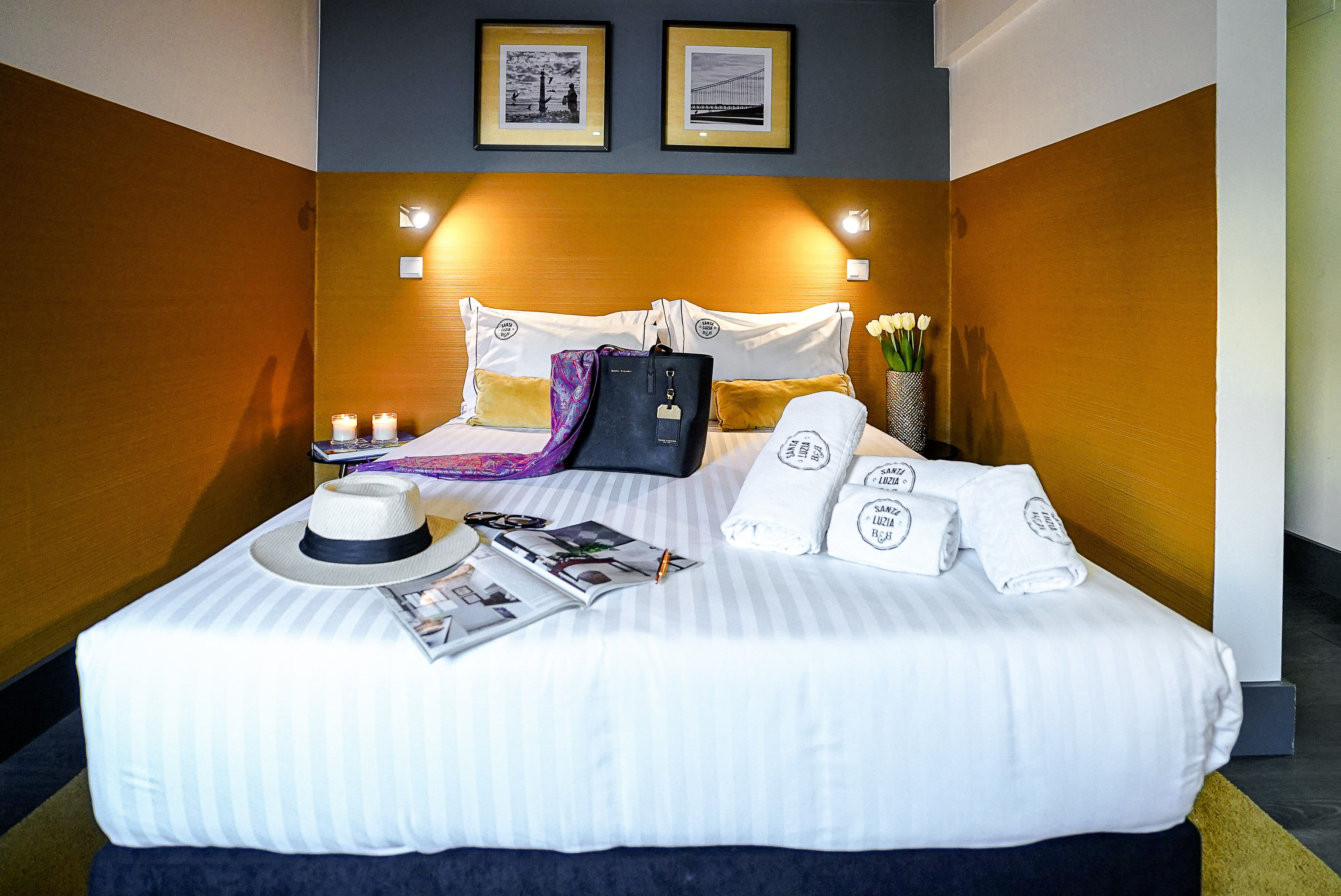 Un lit bien fait, avec du linge de lit blanc, un coussin à rayures, un chapeau, un magazine, un sac noir et des serviettes ornées d’un logo.