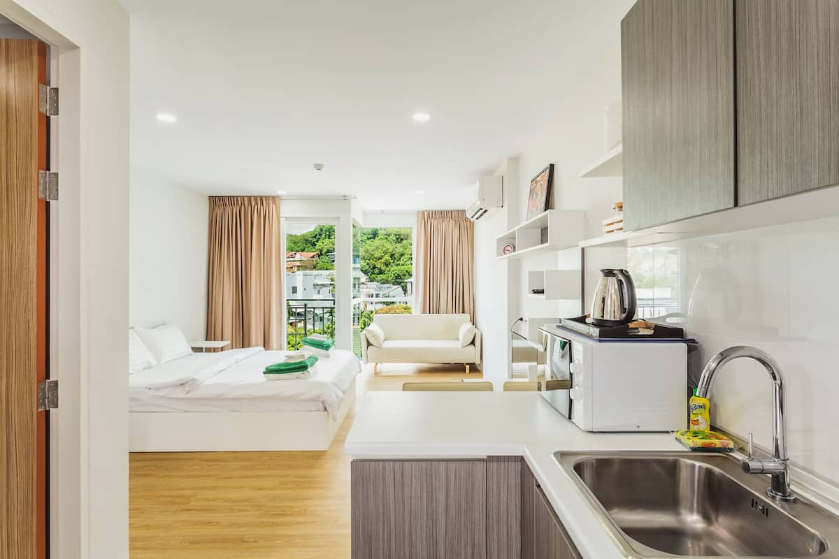 A compact hotel room with a bed, kitchenette, and a view of greenery.