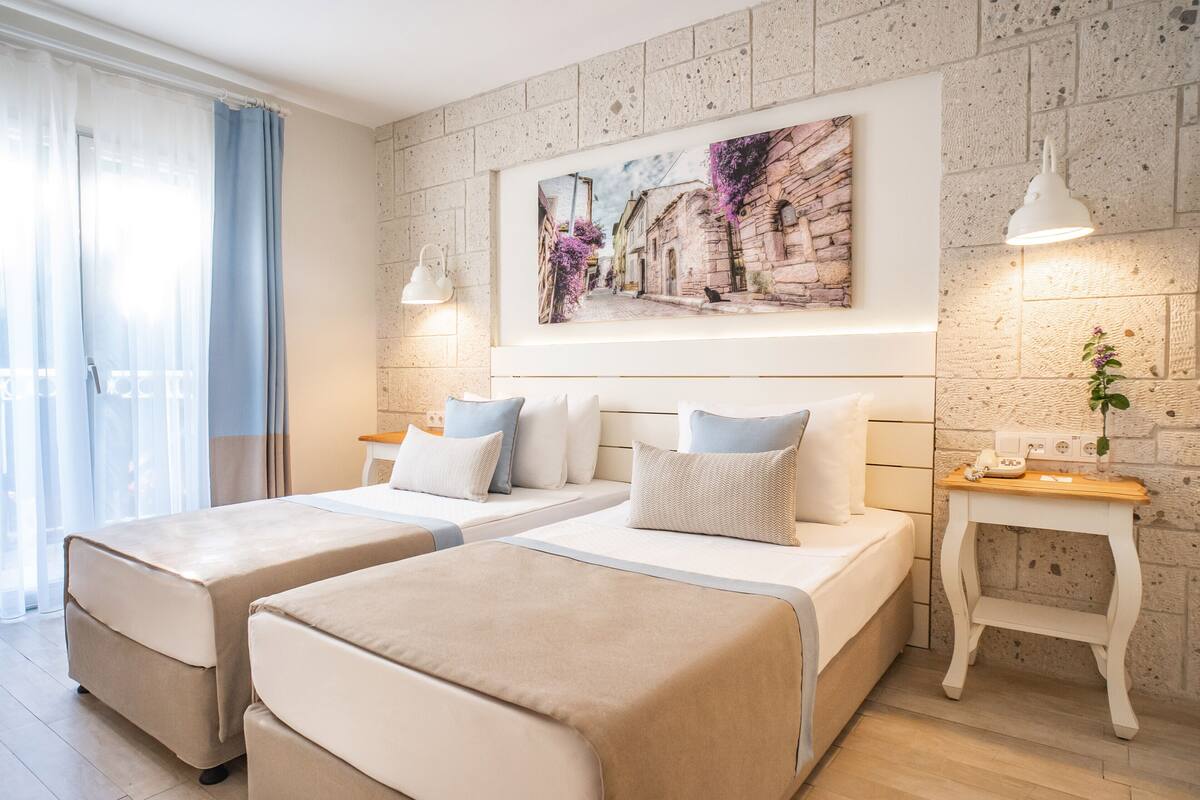 Ein Hotelzimmer mit zwei Betten, einem großen Fenster mit Vorhängen, einem Wandbild, einem Nachttisch mit Lampe und einer kleinen Pflanze.