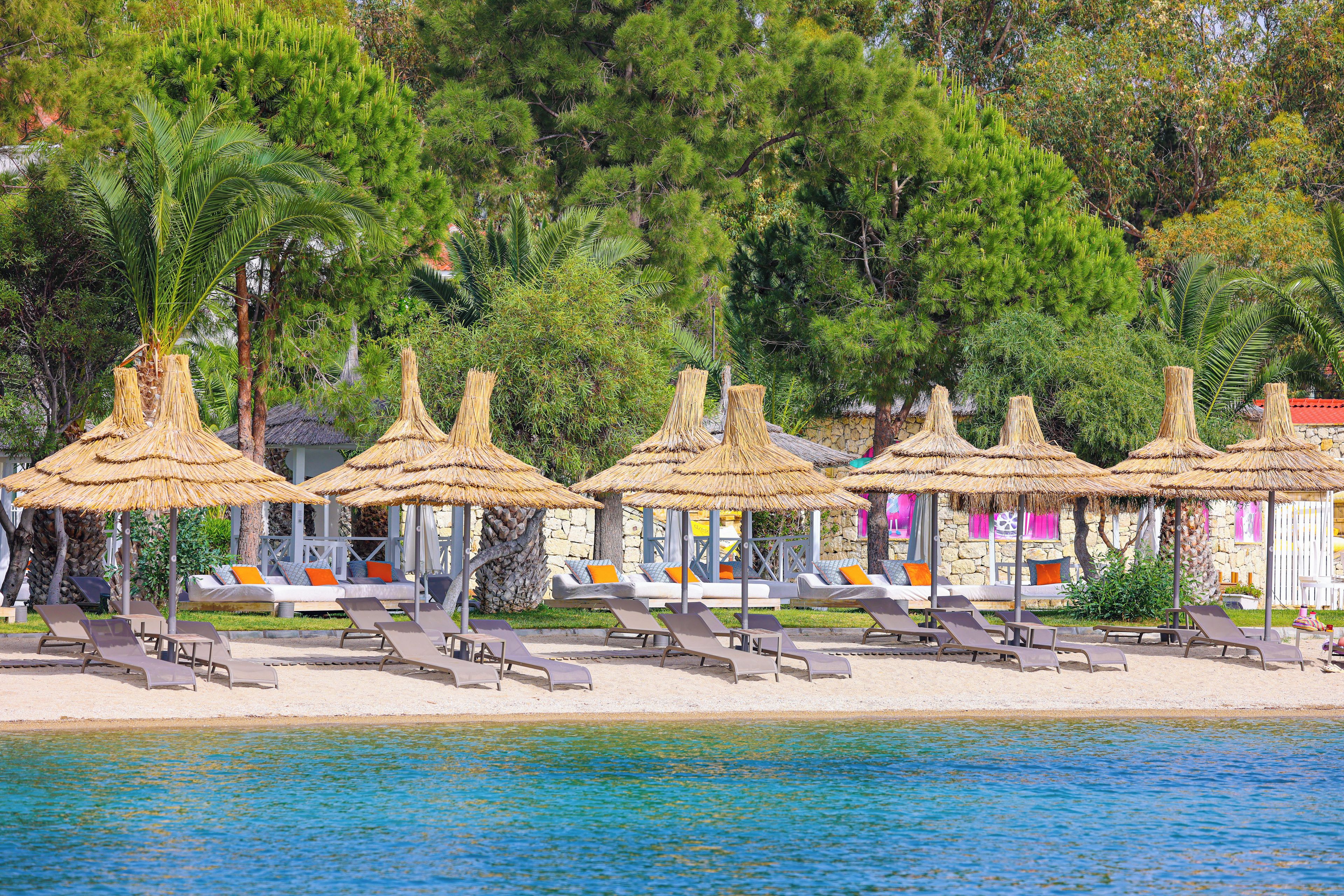 Un resort frente a la playa con sombrillas de paja, sillas de descanso y un edificio de piedra con cortinas coloridas.