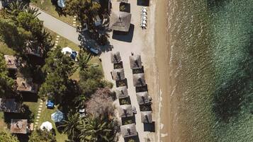 Vista aérea de un resort frente a la playa, con hileras de sombrillas y sillas de tomar el sol, un camino que conduce a la playa y un pequeño edificio con techo rojo.