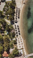Vista aérea de un resort frente a la playa, con hileras de sombrillas y sillas de tomar el sol, un camino que conduce a la playa y un pequeño edificio con techo rojo.