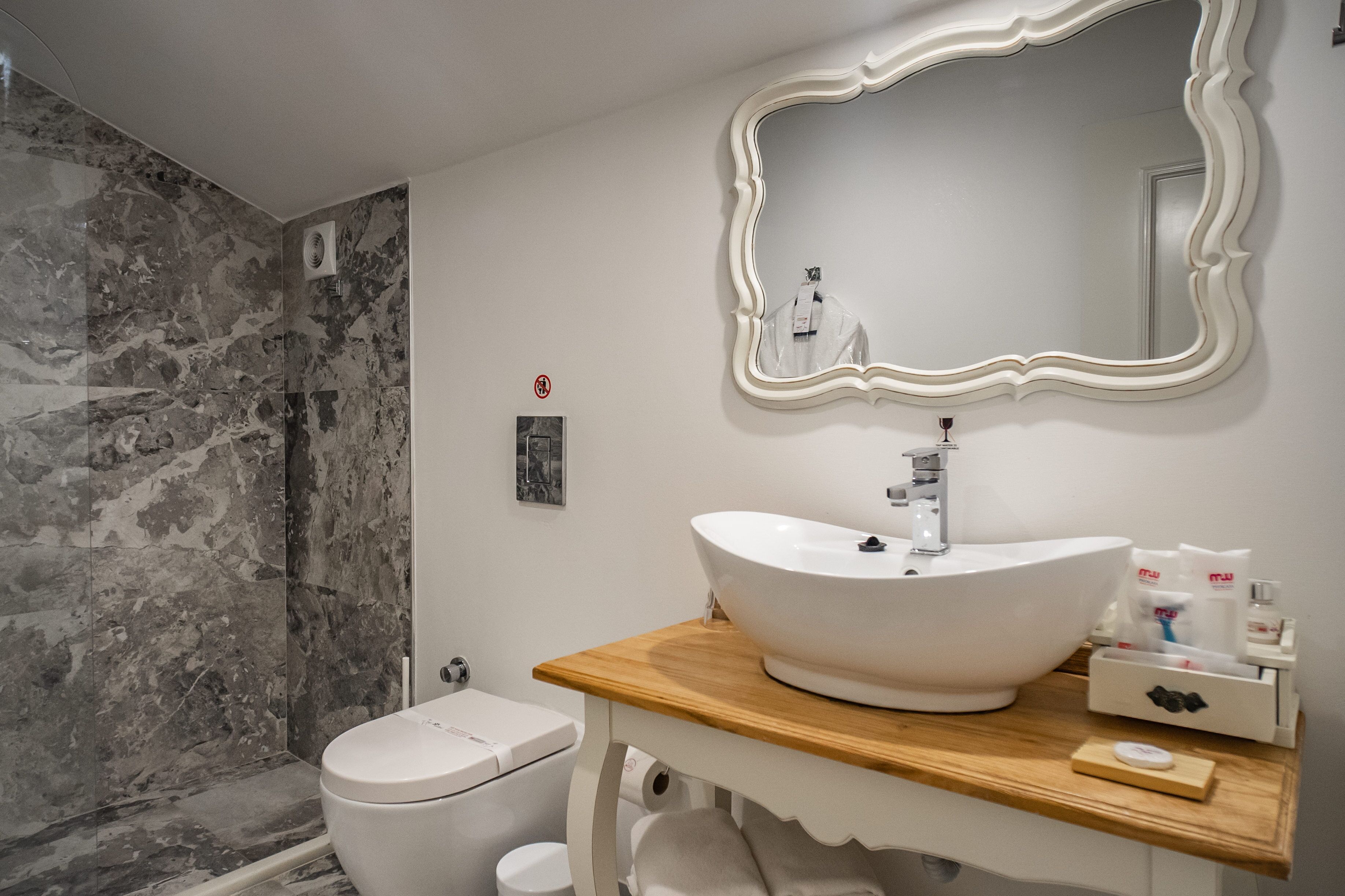 A bathroom with a marble shower wall, a white sink with a wooden countertop, and a large mirror with an ornate frame.