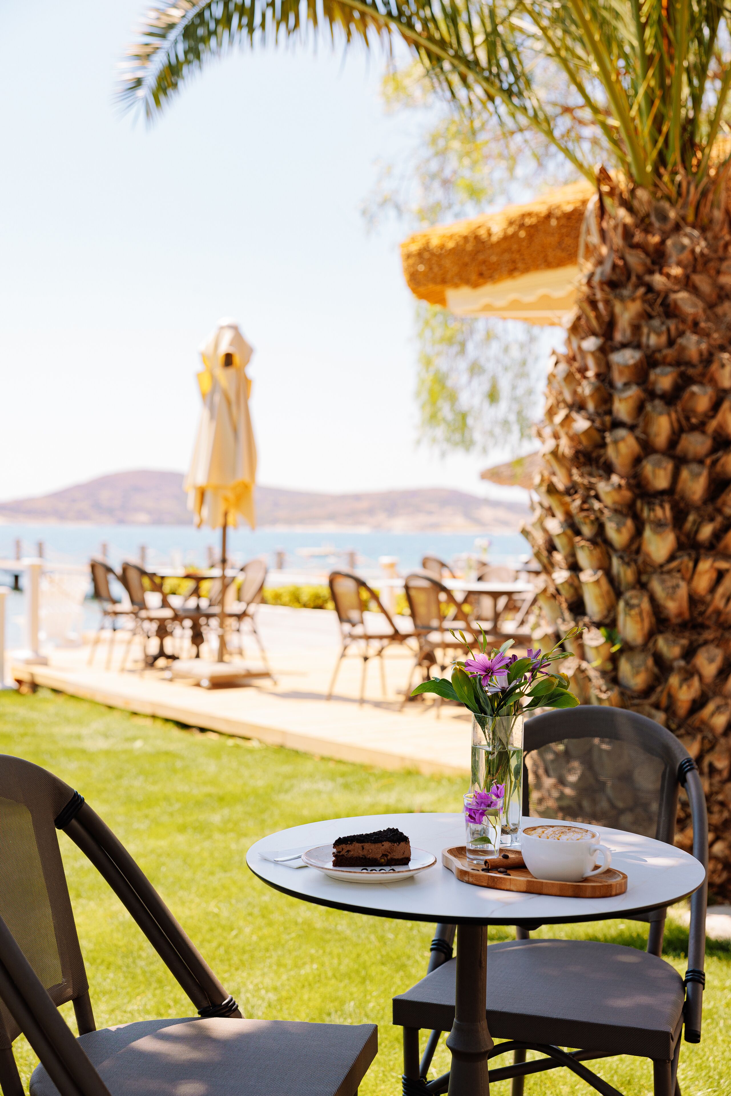 Un área de comedor al aire libre con una mesa para dos, que incluye un postre y una taza de café, con vista a un cuerpo de agua y un paisaje montañoso.