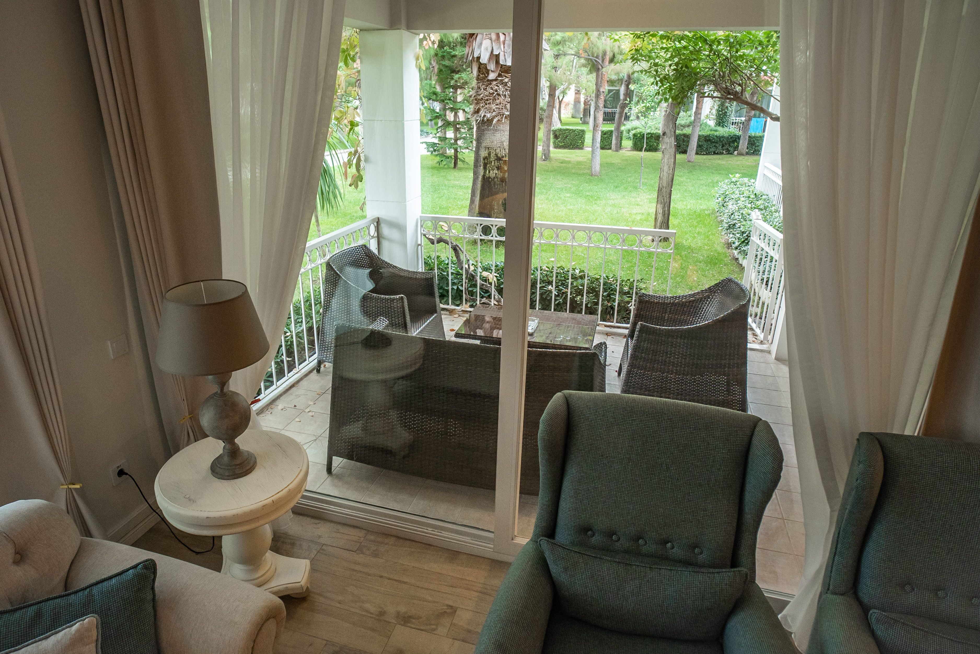 A room with a view of a garden, a dining table with chairs, a lamp, and a sofa.