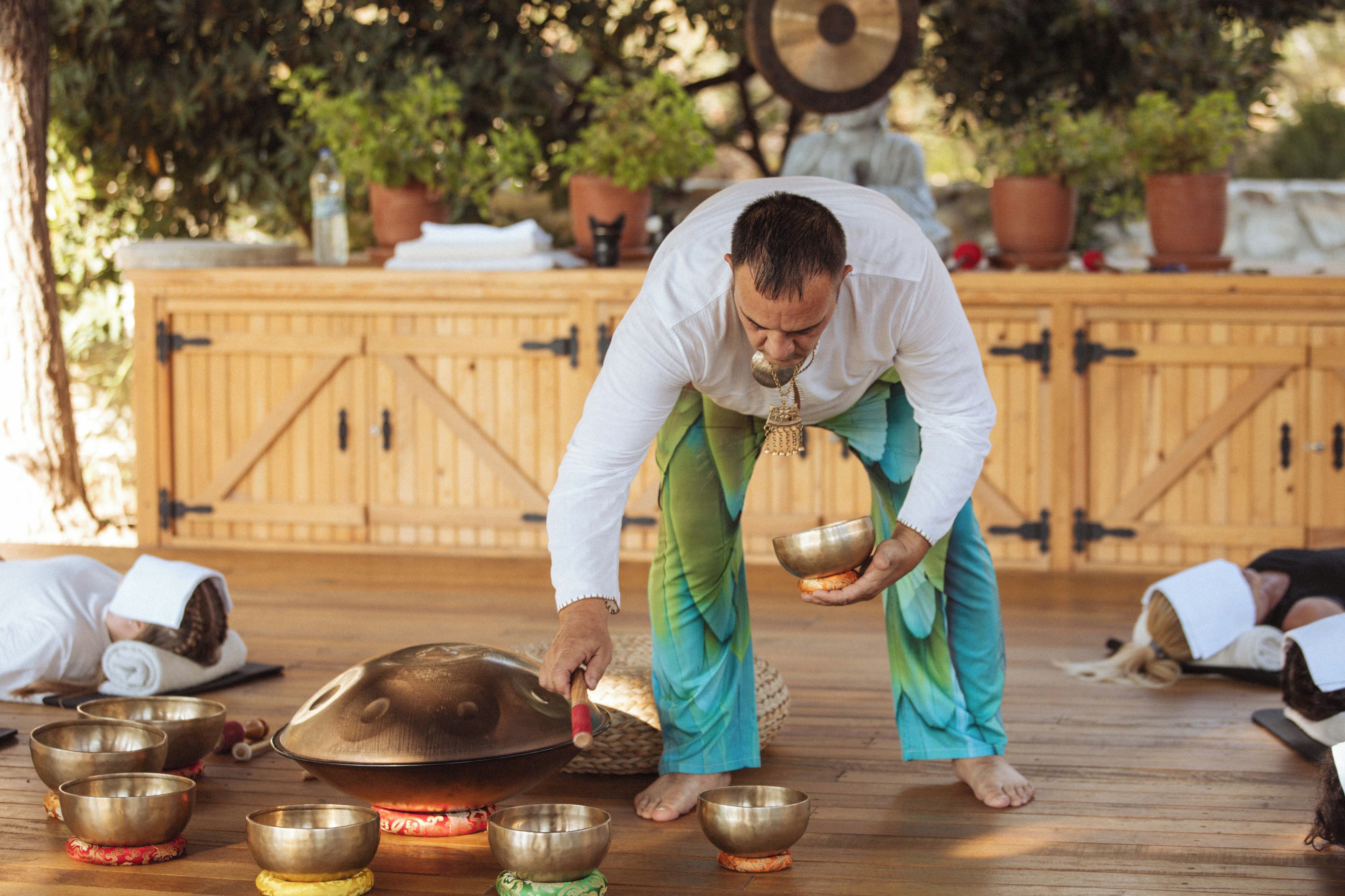 Una persona realiza un ritual con cuencos y una campana en un entorno exterior con estructuras de madera y plantas en macetas.