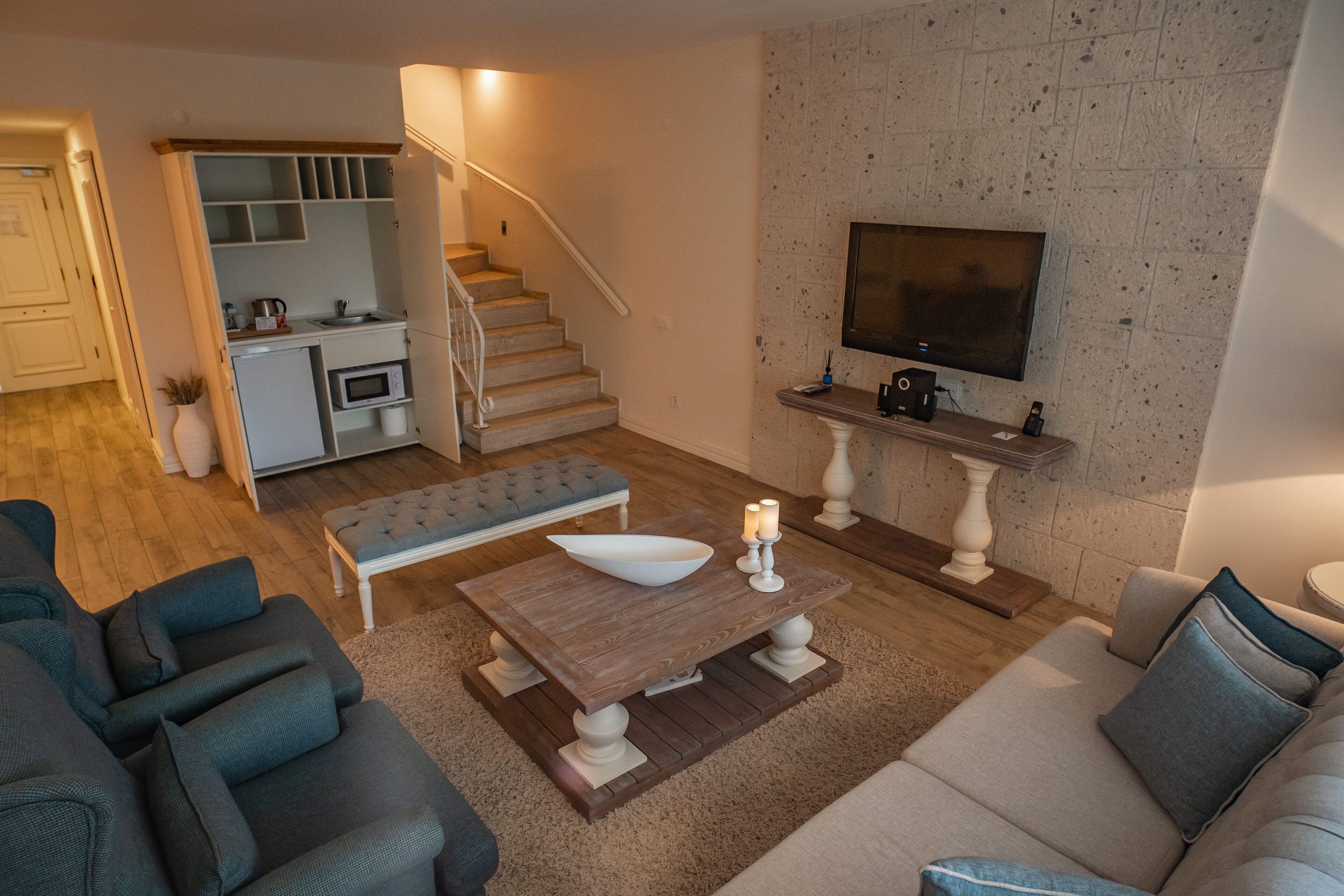Un espacio habitable compacto con kitchenette, una escalera, una zona de comedor con mesa y banco, y un televisor montado en una pared texturizada.