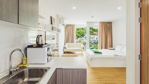 A modern hotel room with a kitchenette, a sofa, a bed, and a view of greenery. - Condo in Kata in Ozone - Unit A606 (Karon)