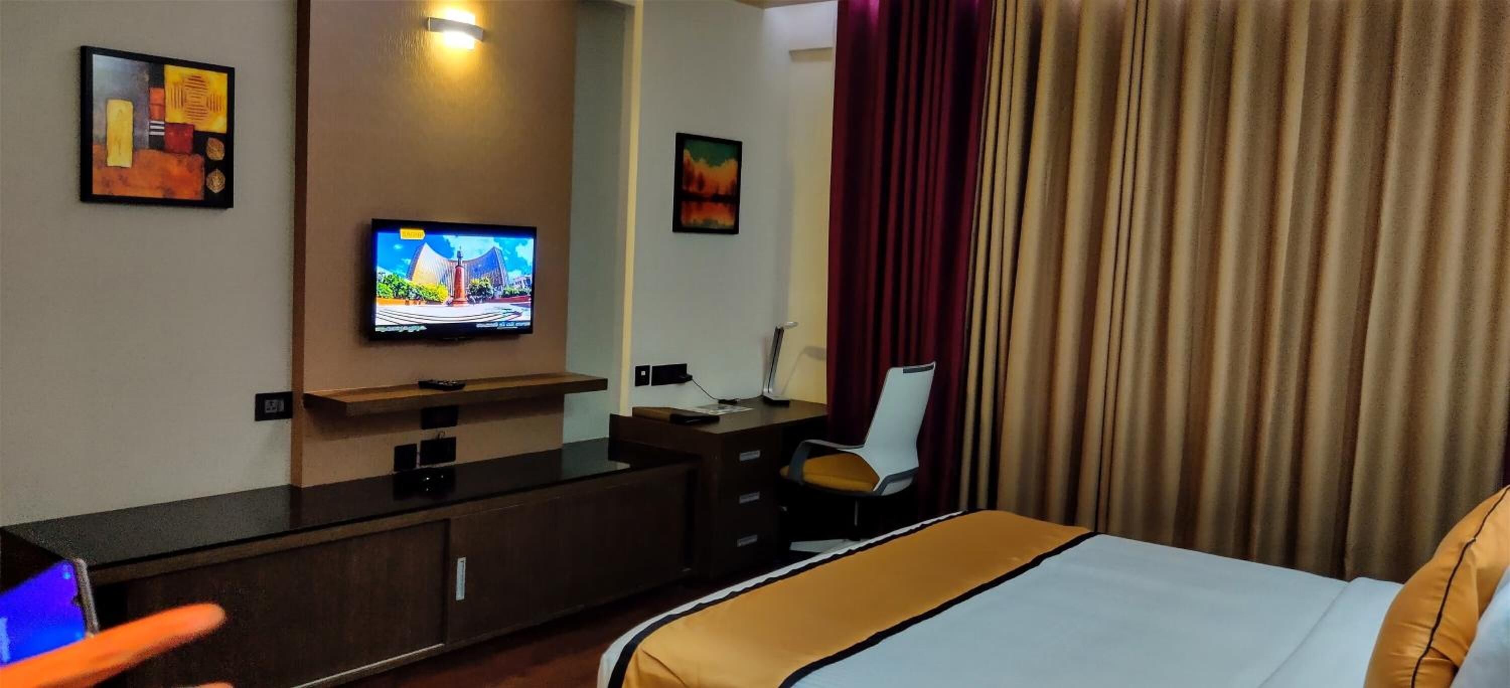 Una habitación de hotel con cama, televisión montada en la pared, escritorio con silla y un amplio ventanal con cortinas.