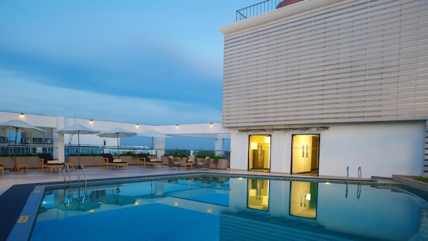 Piscina no terraço com espreguiçadeiras e guarda-sóis, um edifício moderno com grandes janelas e um céu claro.