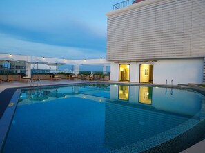 A rooftop swimming pool with lounge chairs and umbrellas, a modern building with large windows, and a clear sky.
