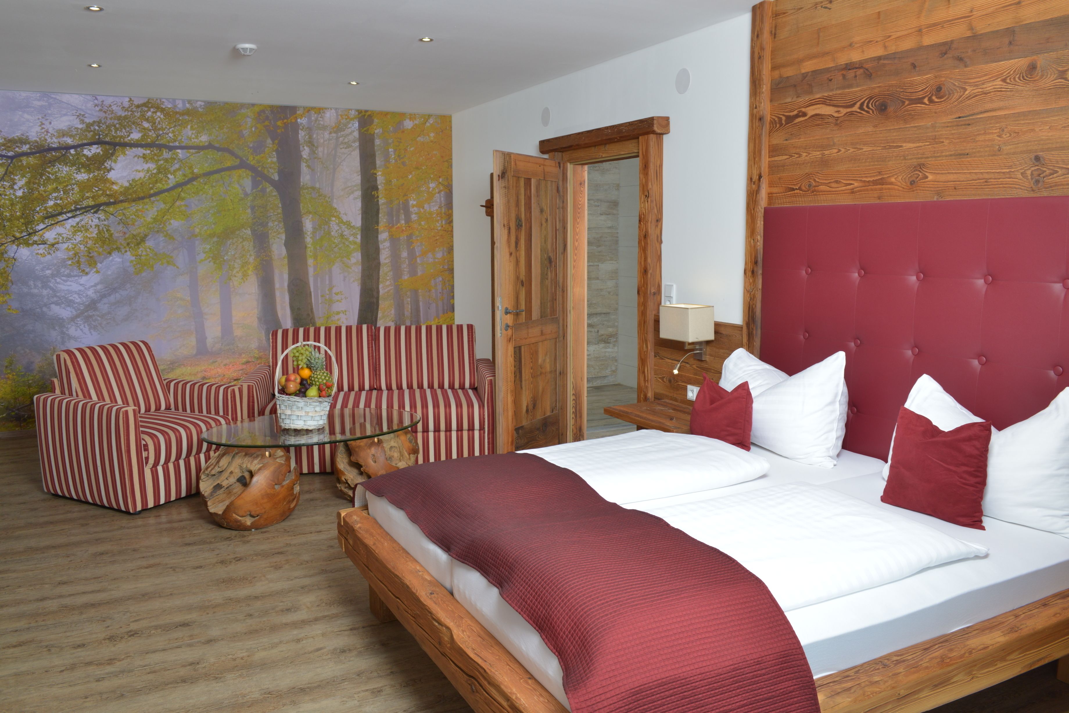 A bedroom with a large bed, striped armchairs, a glass coffee table, and a forest-themed wall mural.