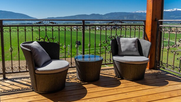 A balcony with two wicker chairs and a small table, overlooking a green field and distant mountains. - Hotel Restaurant Perschler (Fohnsdorf)