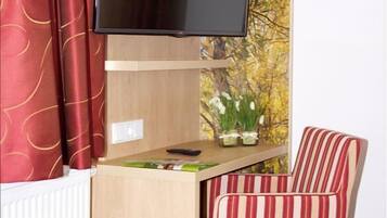 A compact room with a wooden desk, a television mounted on the wall, a striped chair, and a window with curtains.