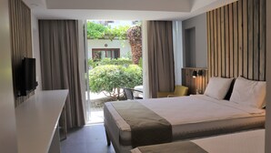 Habitación de hotel con una cama grande, un escritorio y un televisor. Se observa un jardín y un edificio desde la ventana.