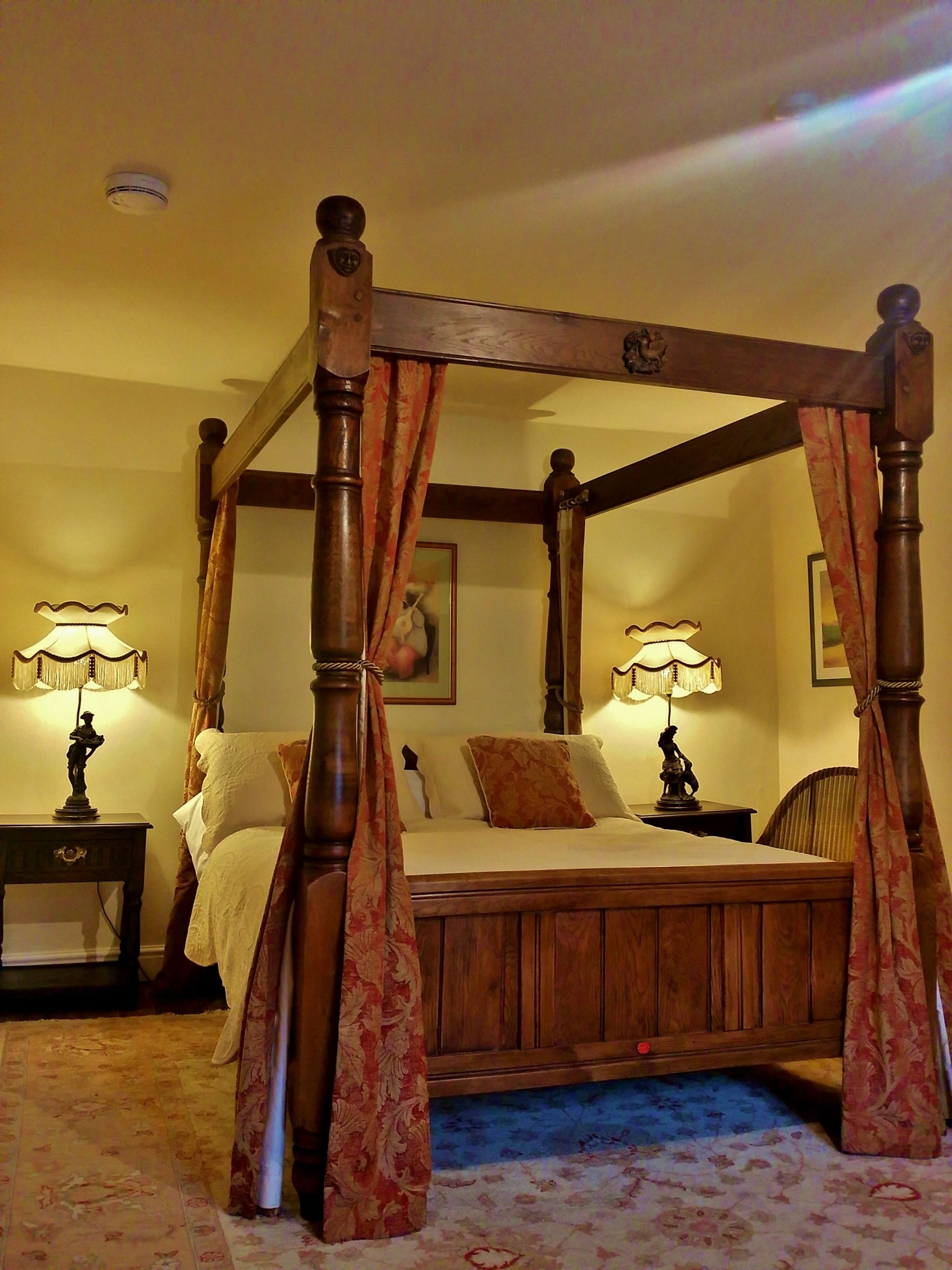 A four-poster canopy bed with a wooden frame and floral curtains, flanked by two bedside tables with lamps, in a room with a carpeted floor.