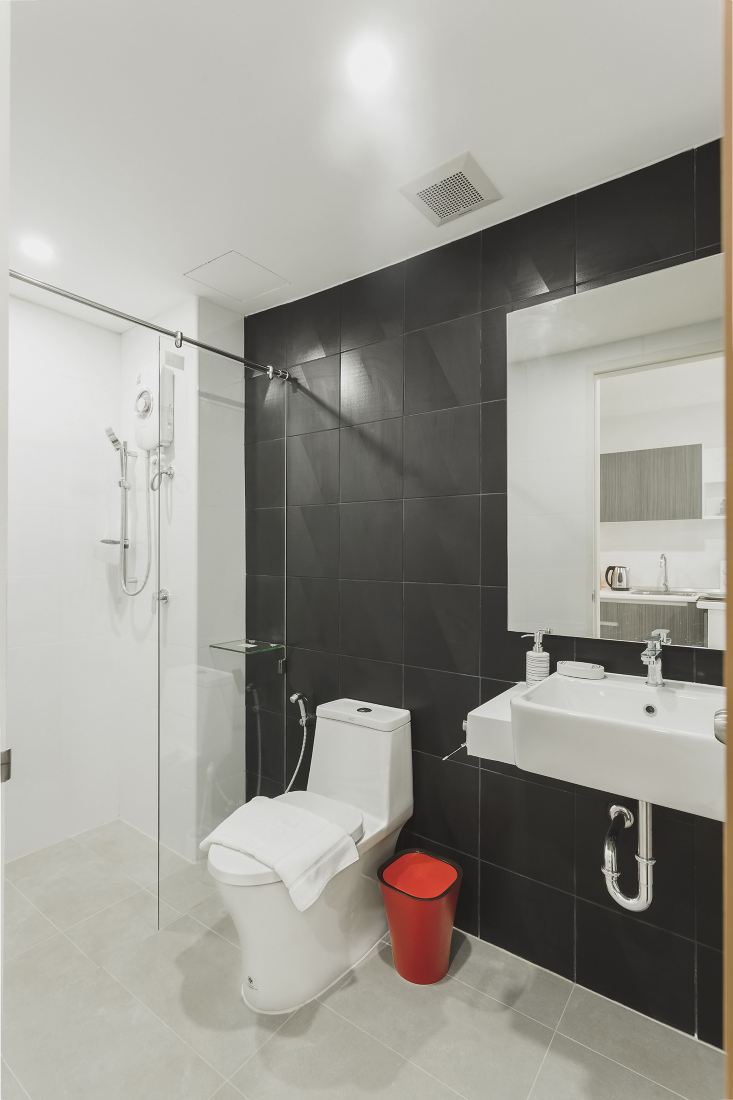 Ein modernes Badezimmer mit einer Duschkabine aus Glas, einer weißen Toilette, einer roten Mülltonne und einem weißen Waschbecken mit Spiegel darüber.