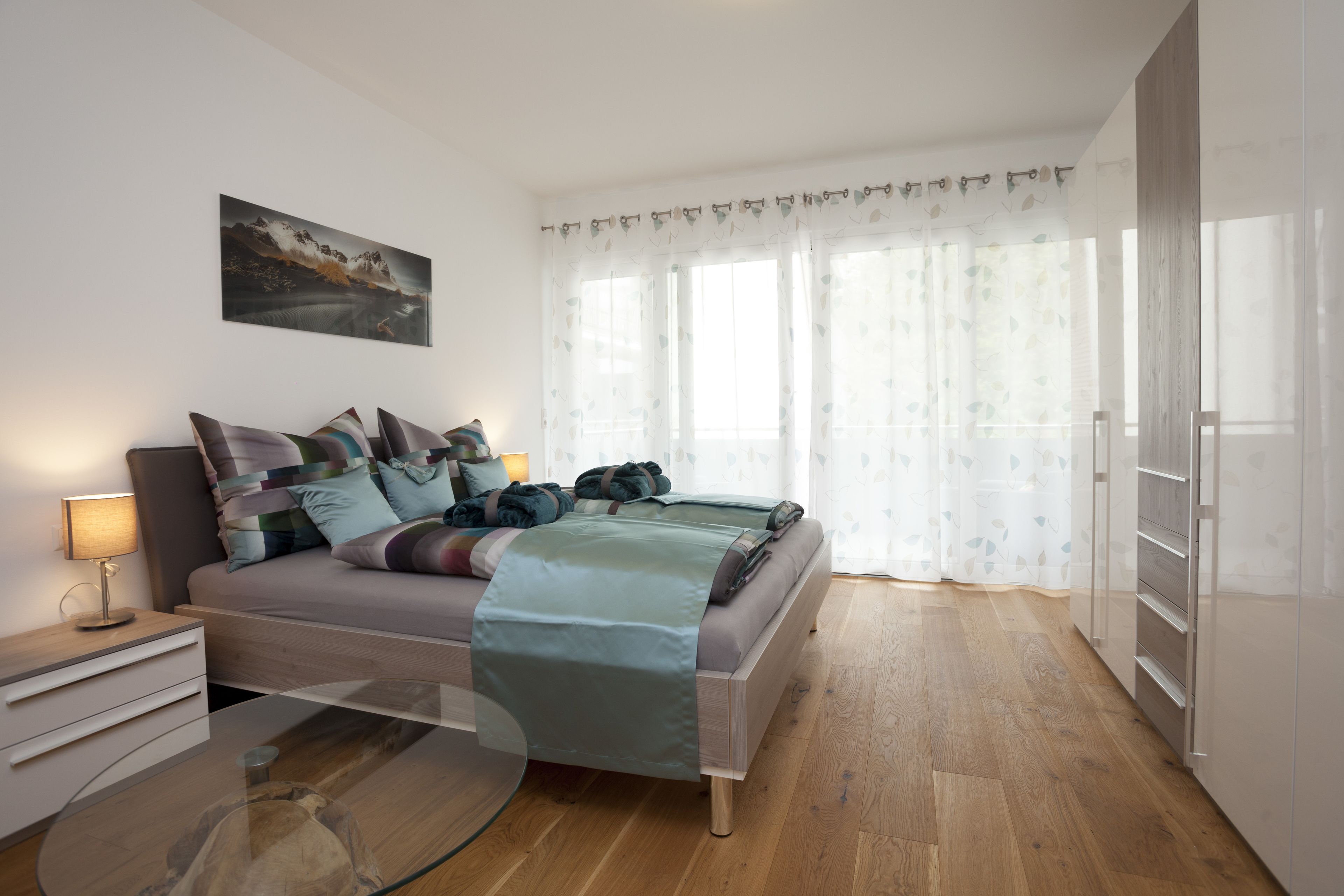 A modern bedroom with a bed, bedside tables, a glass coffee table, and a painting on the wall.