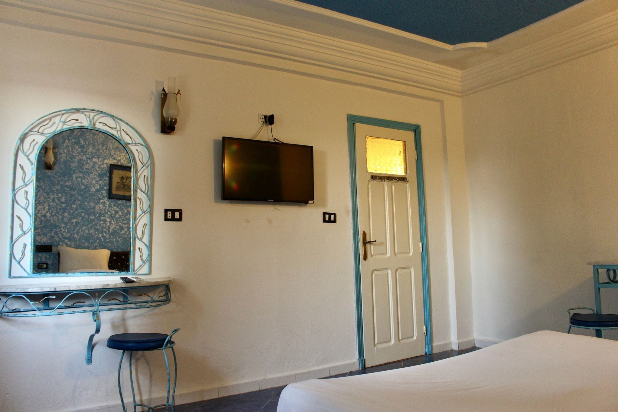 A hotel room with a bed, a TV mounted on the wall, a door with a light above it, and a mirror with a decorative frame.