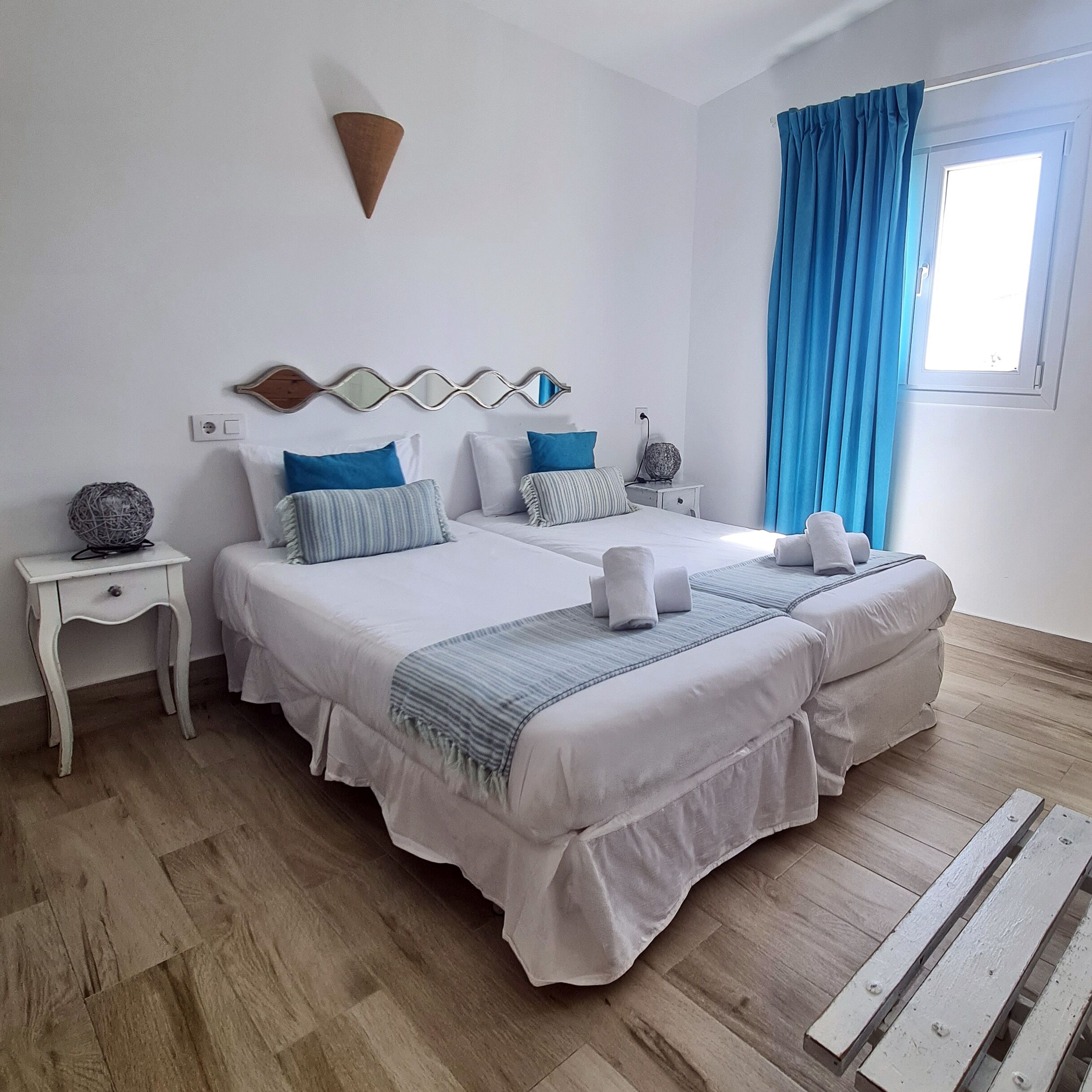 A double bed with white bedding and blue accents, a bedside table with a lamp, and a window with blue curtains.