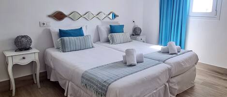 A double bed with white bedding and blue accents, a bedside table with a lamp, and a window with blue curtains.