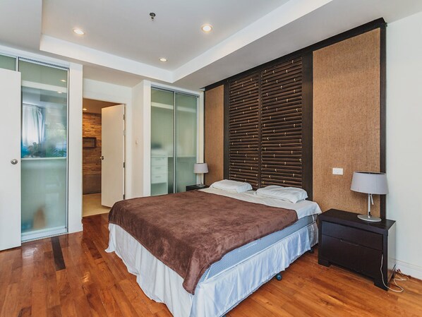 A modern bedroom with a large bed, a wooden headboard, a nightstand with a lamp, and a sliding glass door leading to a bathroom.