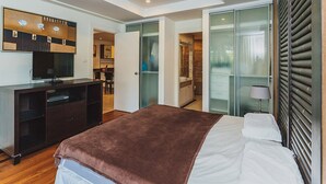 A modern bedroom with a bed, a TV, a desk, and a bathroom visible through a glass door. - Condo in Panwa in Bel Air - Unit C204 (Wichit)