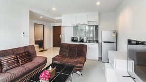 A modern living room with a brown sofa, a coffee table, and a small table with a vase of flowers. A kitchen with white cabinets and appliances is visible in the background. - Condo in Panwa in Pixels 89-230 (Wichit)