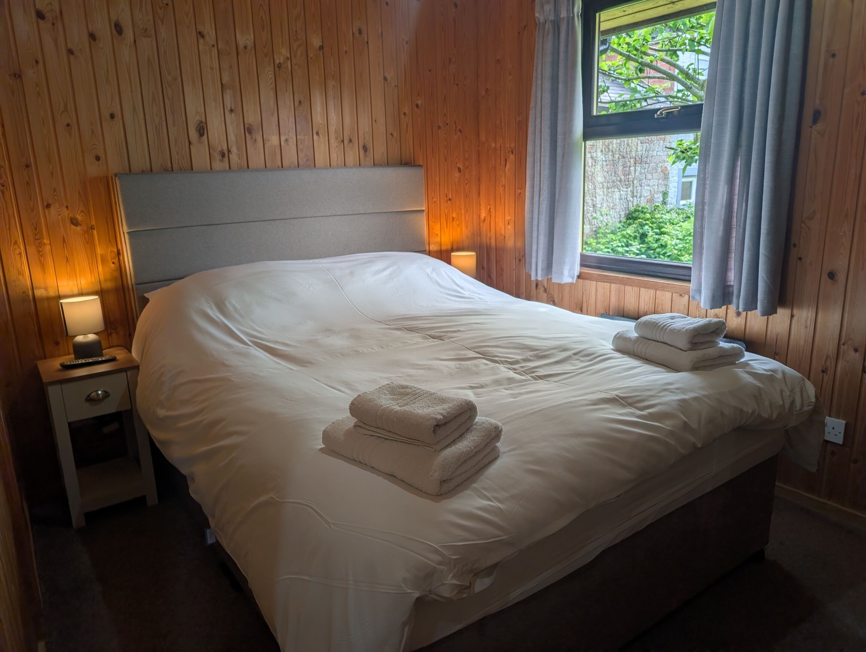 A neatly made bed with white linens and folded towels, a bedside lamp, and a window with curtains.