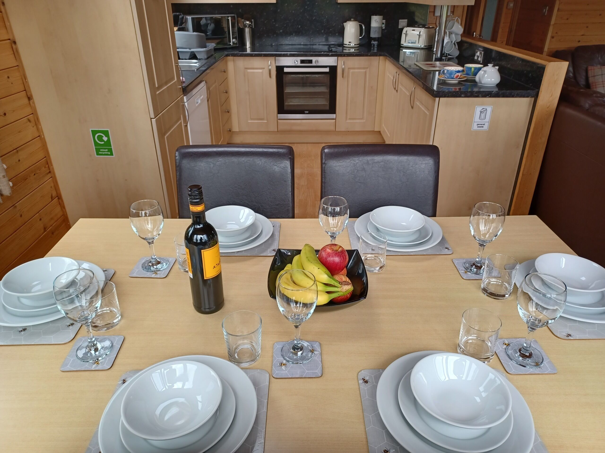 A dining table set with plates, glasses, and a bottle of wine, with a fruit bowl in the center.