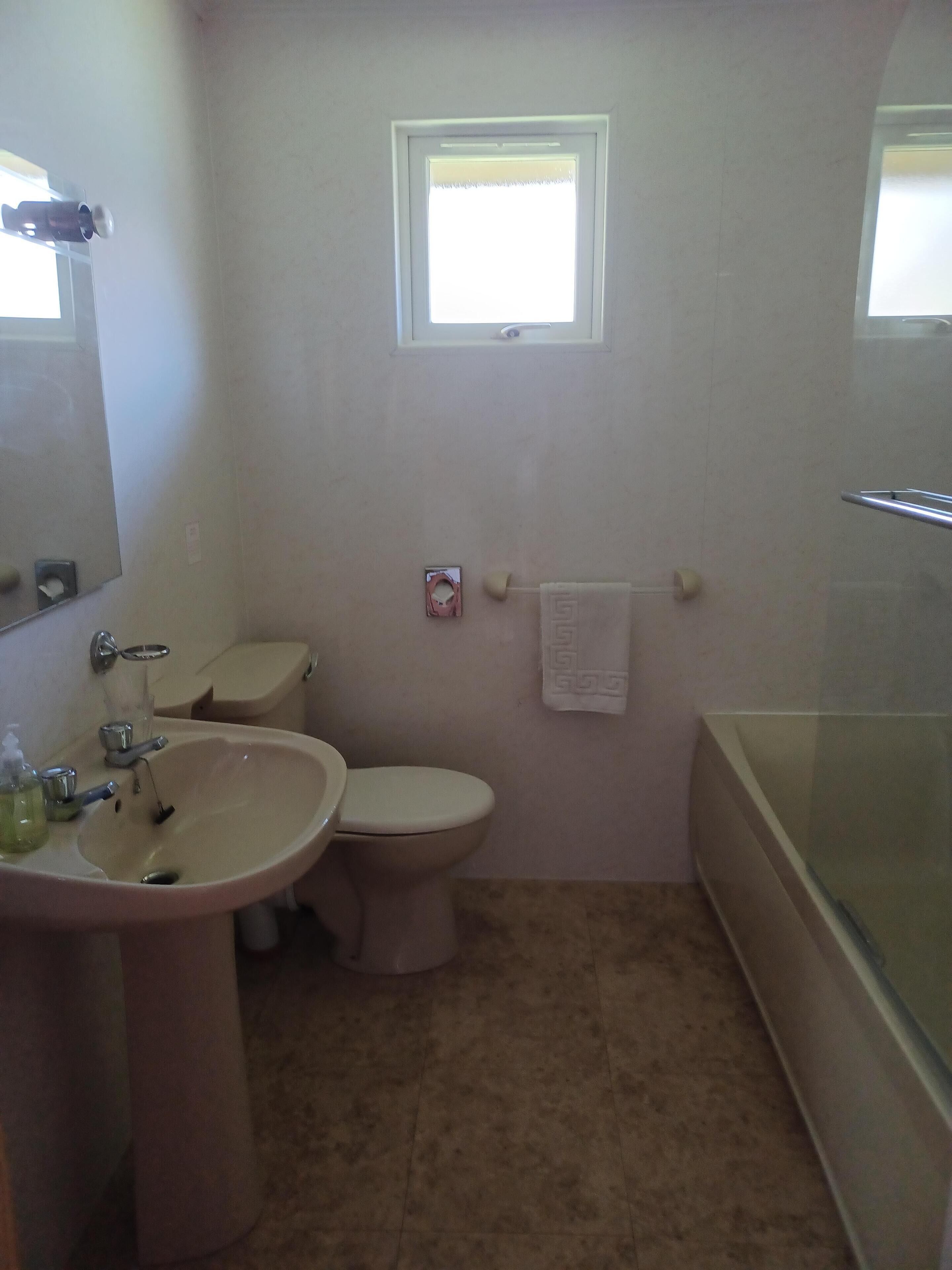 Un baño con lavamanos, inodoro y bañera. Hay una ventana sobre el inodoro y una toalla colgada en la pared.