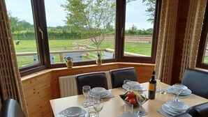 Une table à manger est dressée avec des assiettes, des verres et une bouteille, et offre une vue sur un jardin et une clôture en bois à l’extérieur.