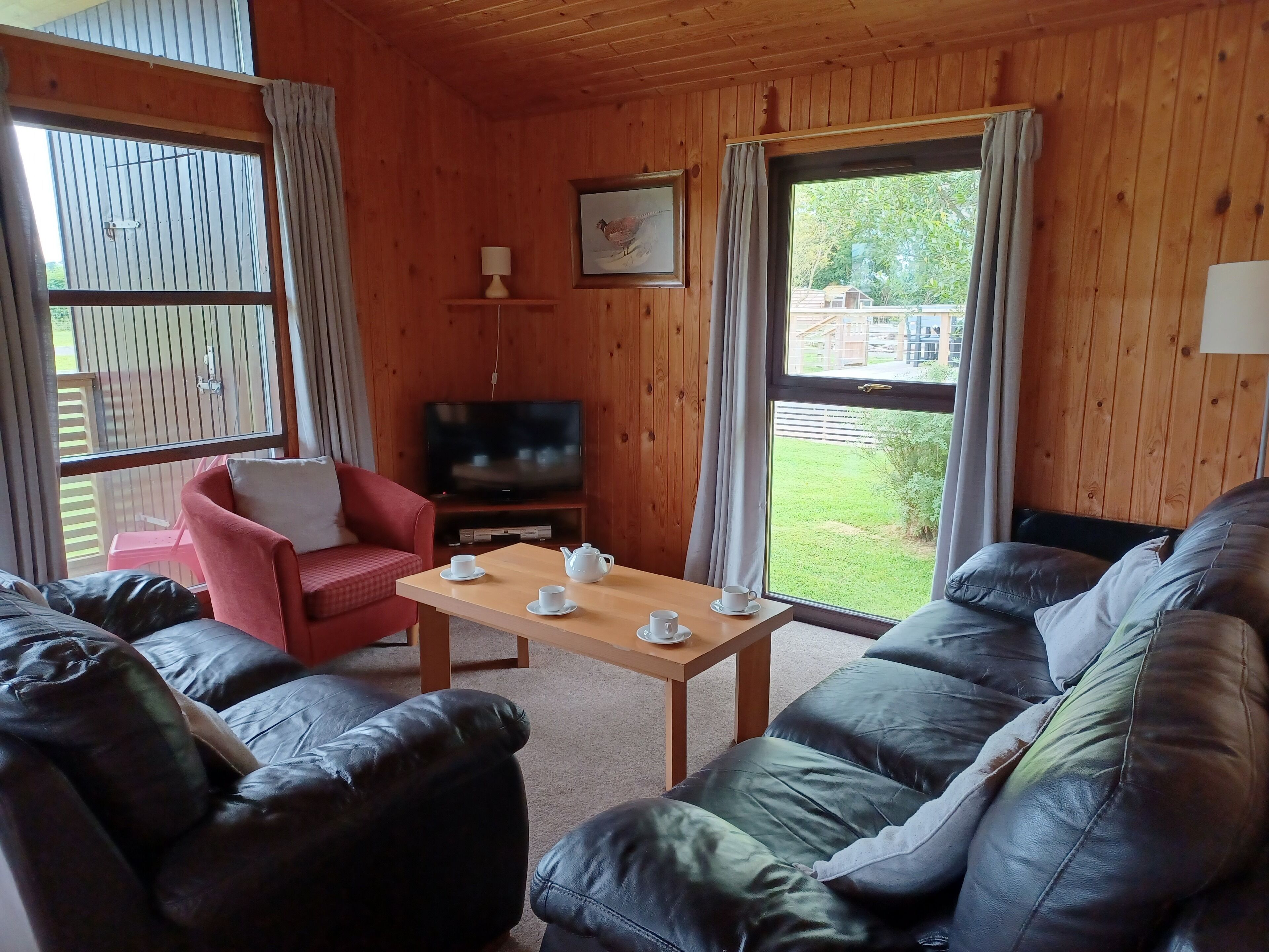 Une pièce chaleureuse avec un décor intérieur en bois, un téléviseur posé sur un meuble, un fauteuil rouge, un canapé en cuir noir et une table avec des tasses et une théière.