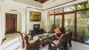 A spacious living room with a high ceiling, a large window with a view of greenery, a wooden coffee table, and a television on a cabinet.