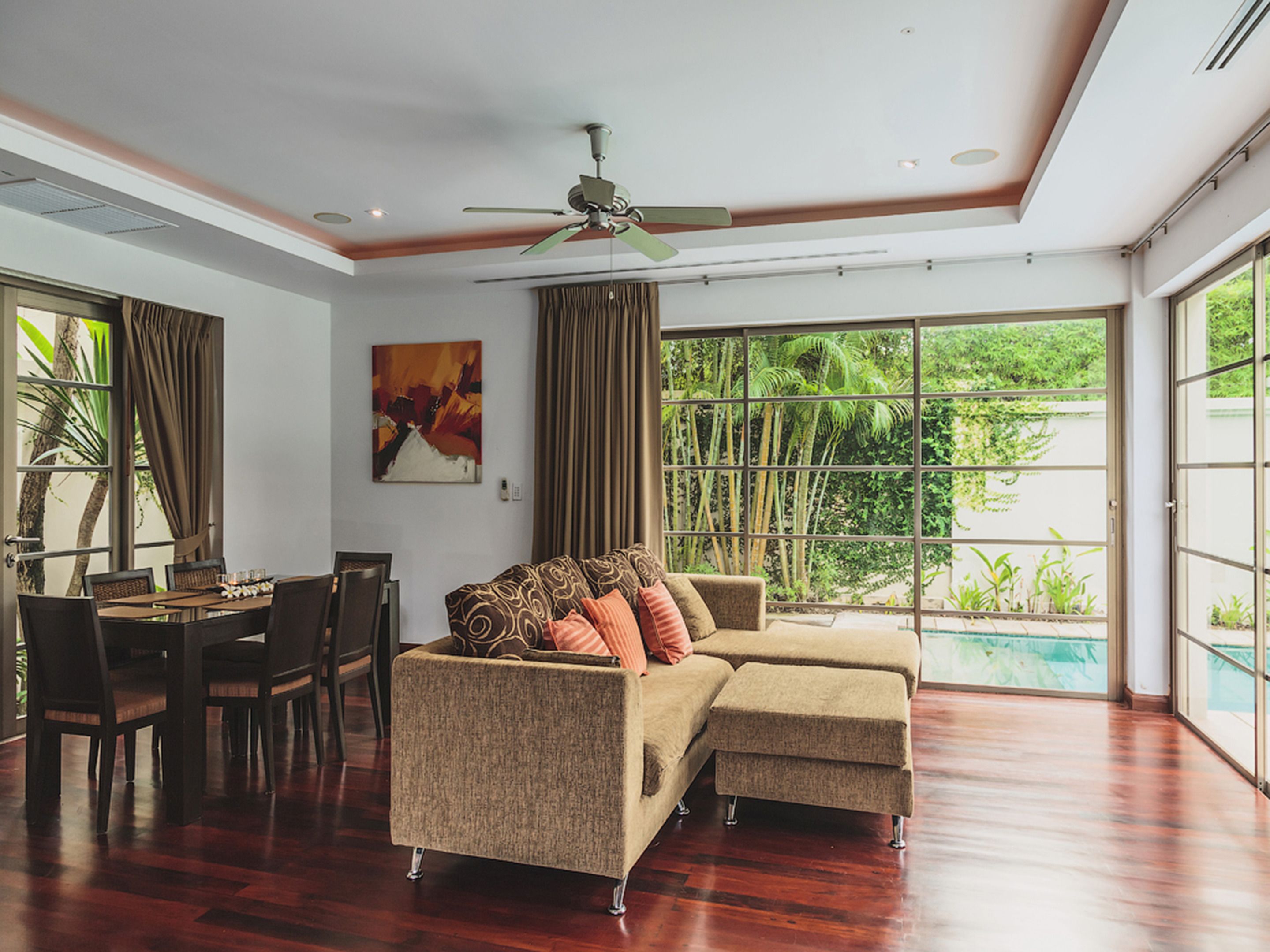 A spacious living area with a large sectional sofa, a dining table, and chairs. There is a ceiling fan, a painting on the wall, and a view of greenery through large windows.