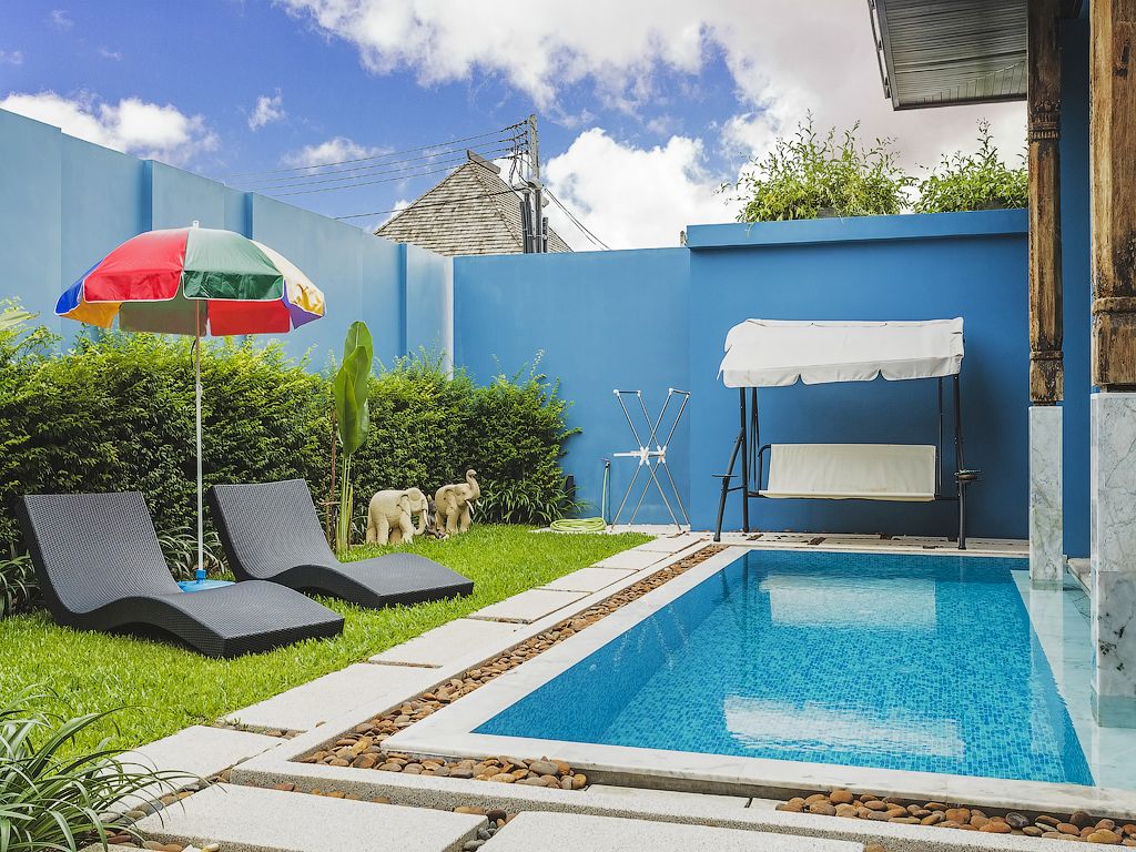A swimming pool with lounge chairs and a covered seating area, surrounded by a blue wall and greenery.