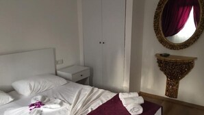 A neatly made bed with white linens and a purple bedspread, a white wardrobe, a small bedside table, and a decorative mirror with a gold frame. - Cohiba Hotel (Bodrum)