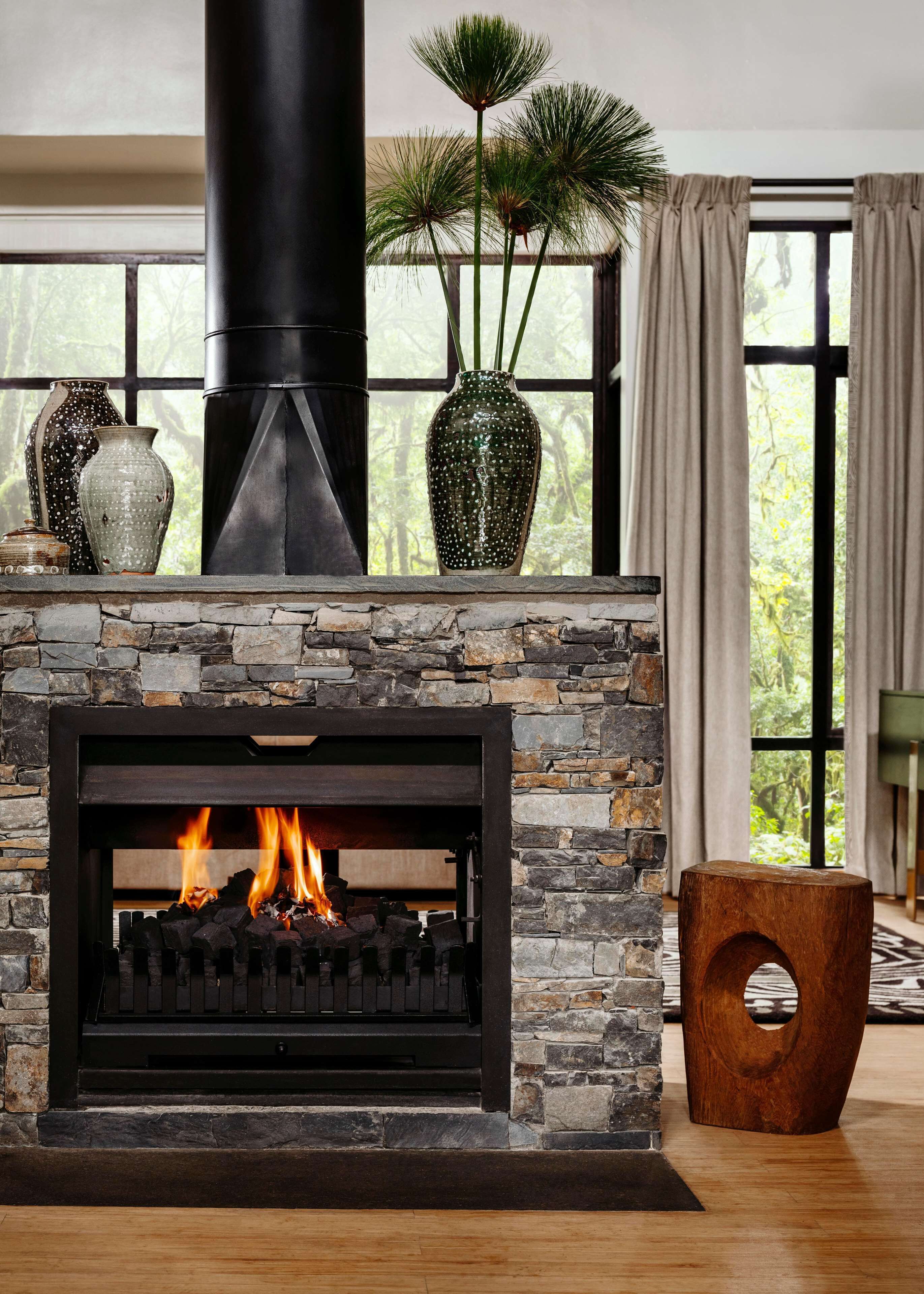 A stone fireplace with a black metal chimney, a fire burning inside, and a window with curtains.