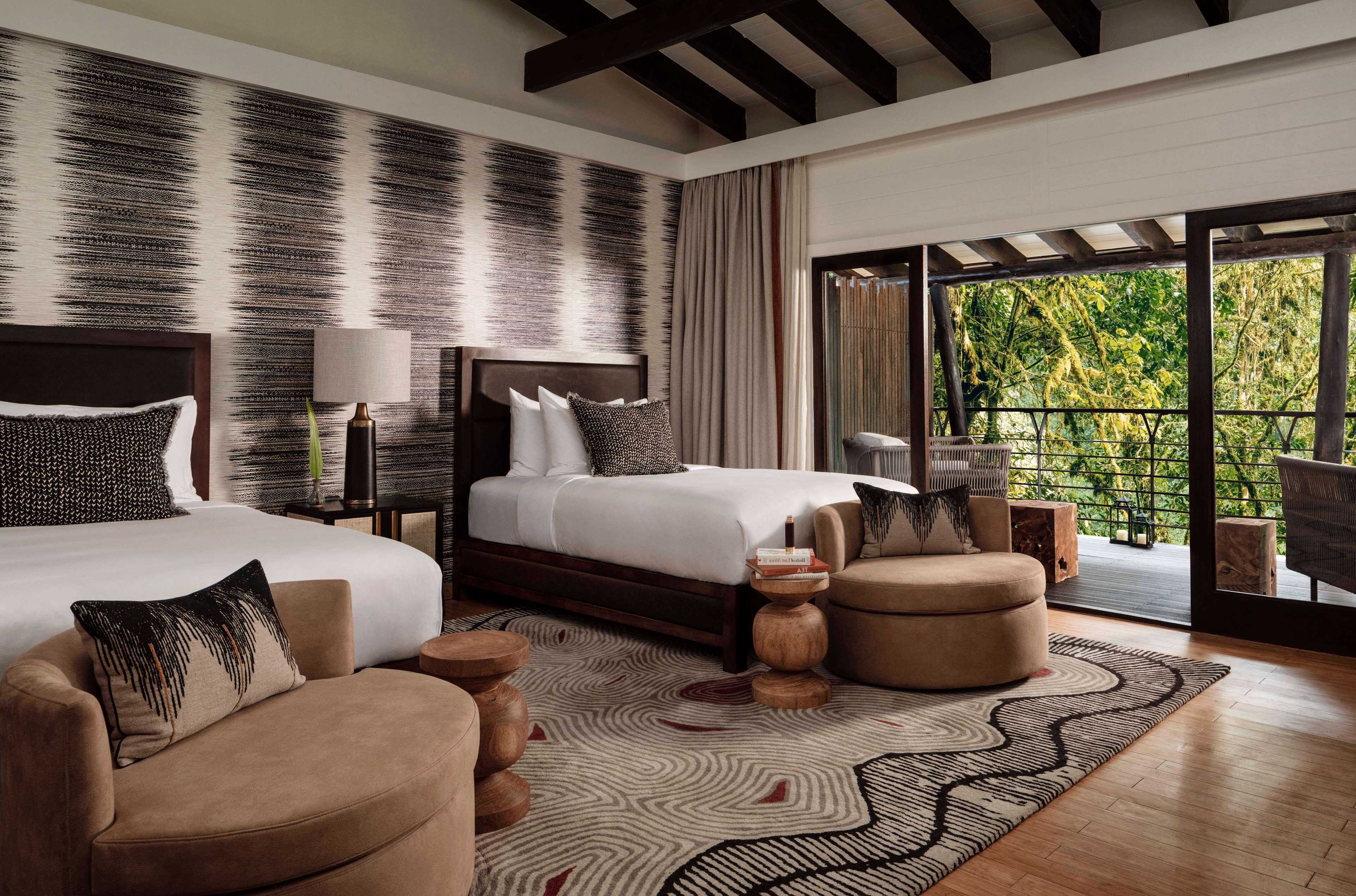 A modern bedroom with a large bed, two bedside tables, a sofa, and a view of greenery through a balcony.