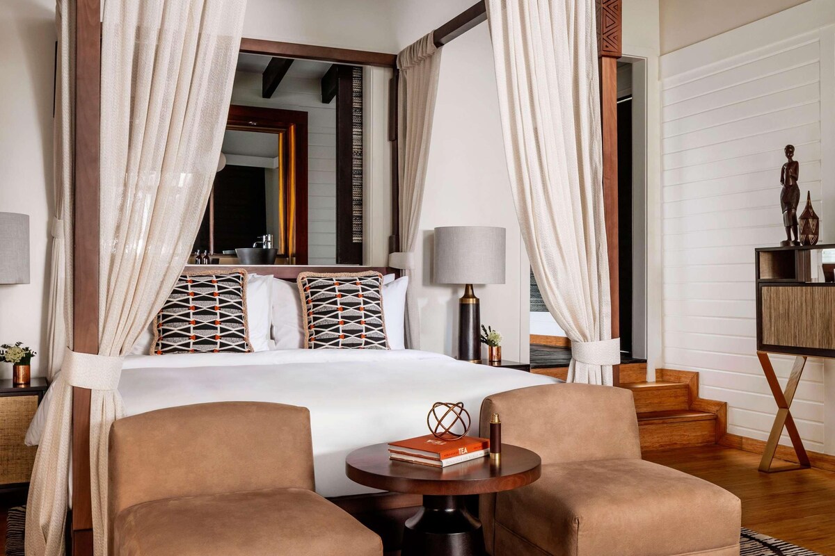 A four-poster bed with white curtains, a round wooden table, two armchairs, and a patterned rug.