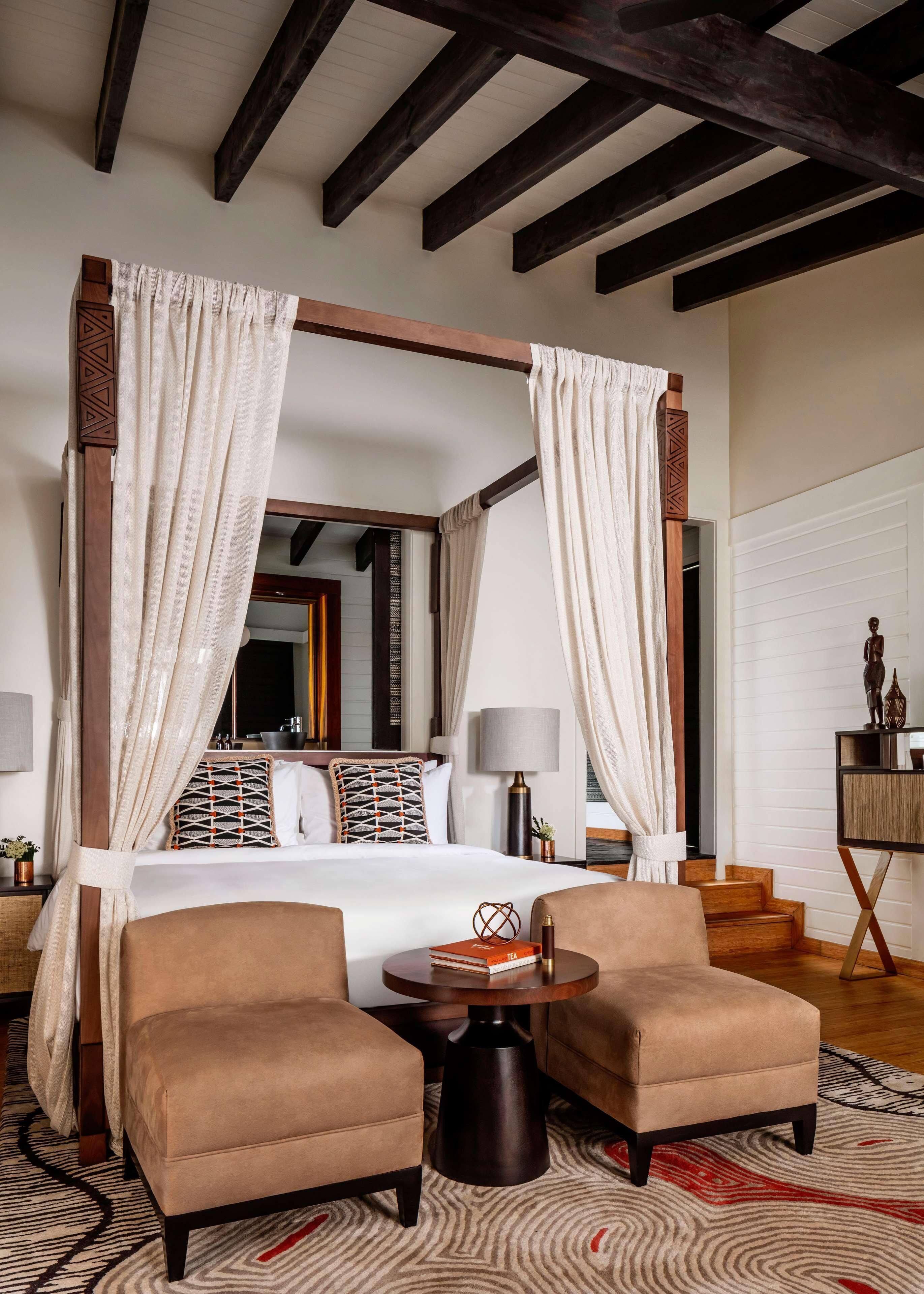 A four-poster bed with white curtains, a round wooden table, two armchairs, and a patterned rug.