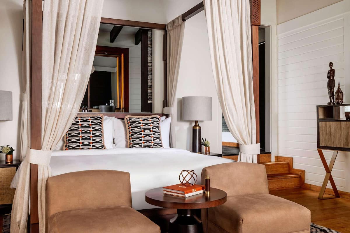 A four-poster bed with white curtains, a round wooden table, two armchairs, and a patterned rug.