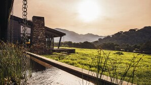 A modern house with a stone facade, a covered patio, and a view of a green field and mountains in the background. - One&Only Nyungwe House (Bushekeri)