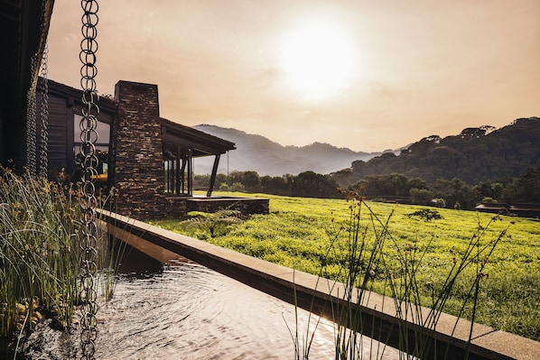A modern house with a stone facade, a covered patio, and a view of a green field and mountains in the background. - One&Only Nyungwe House (Bushekeri)