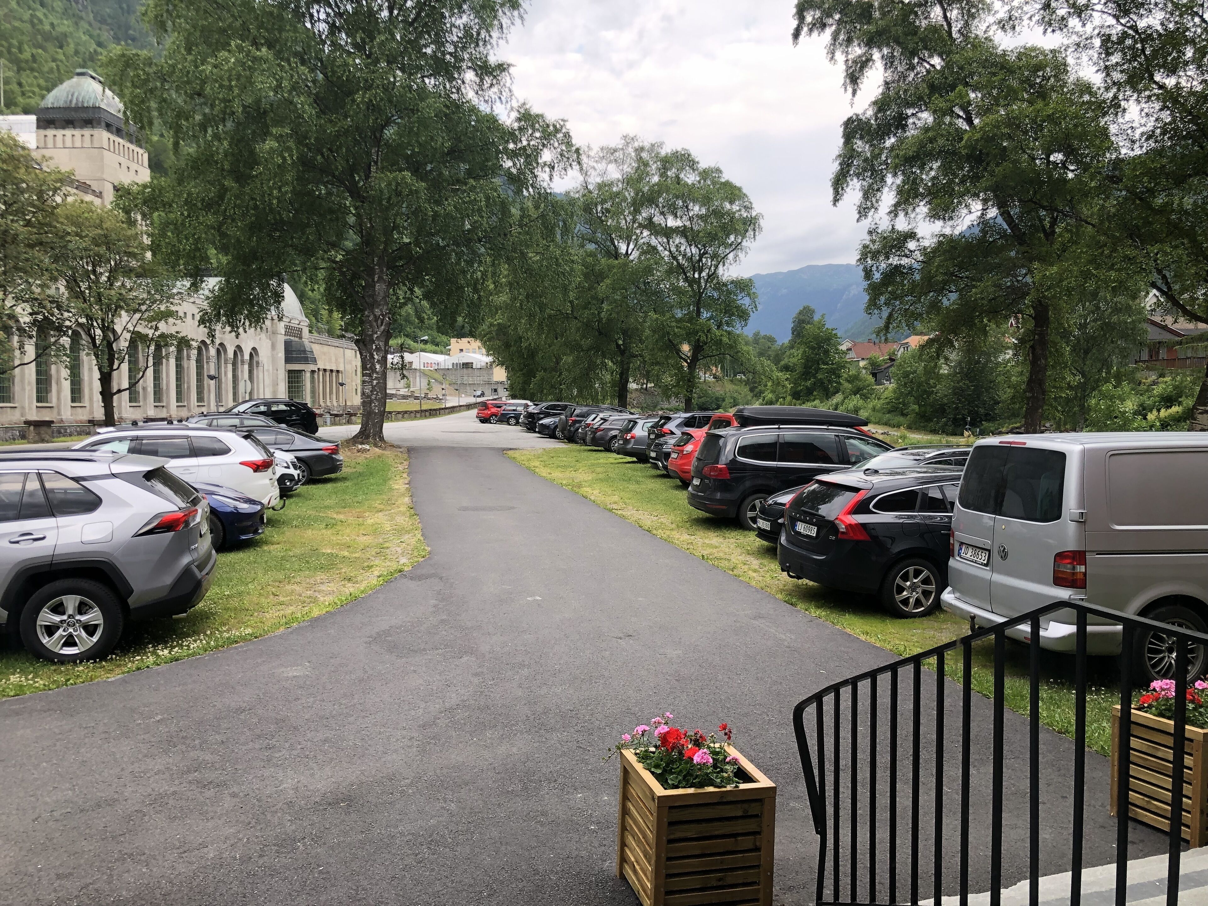 A parking area with cars parked along a path, surrounded by trees and a building with a dome in the background.