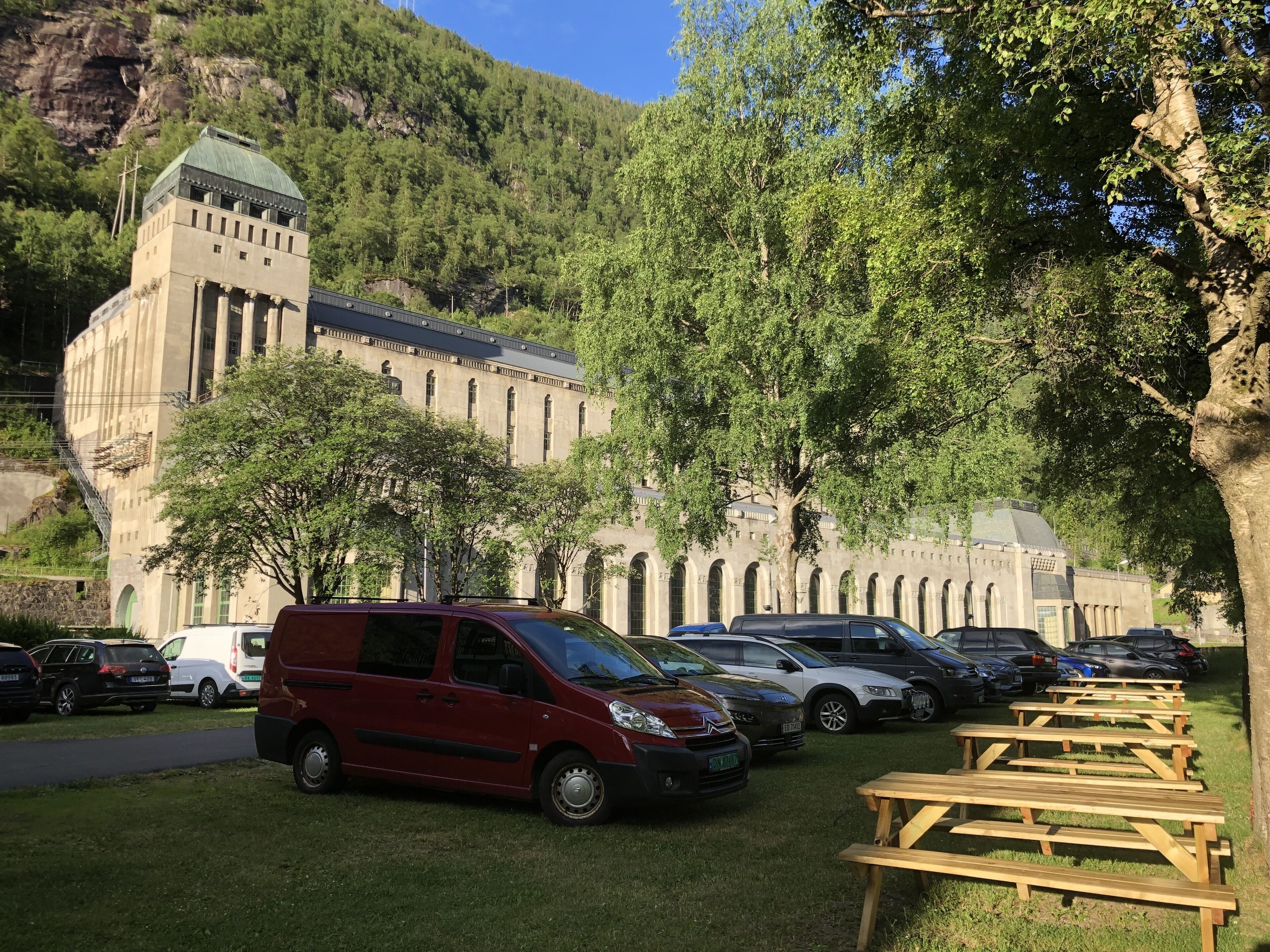 Un estacionamiento con automóviles y mesas de picnic frente a un edificio con una torre y ventanas arqueadas.